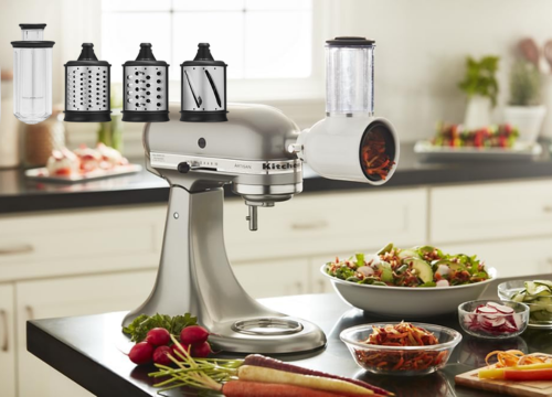  The image displays a KitchenAid 5KSMVSA Fresh Prep Slicer/Shredder attached to a silver KitchenAid stand mixer. The slicer/shredder is white with a transparent chamber, and it's shown with four different attachments. The setting is a kitchen counter with various sliced and shredded vegetables, including carrots, radishes, and a salad, suggesting the appliance's use in food preparation.