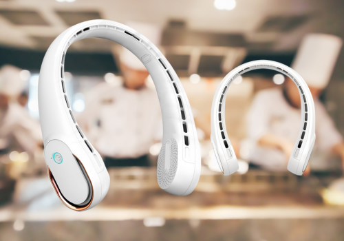 Two white portable neck fans are floating in the foreground, angled slightly differently to show their design. The background is blurred, but it appears to be a kitchen or food preparation area with individuals wearing white chef hats.