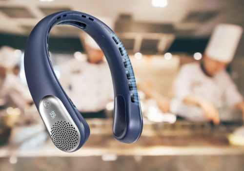 A navy blue portable neck fan is featured in the foreground. The background is blurred, showing individuals in white chef hats working in a kitchen setting. The fan has a digital display showing '99'.