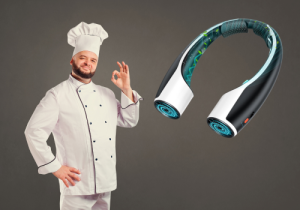 A black and white portable neck fan is featured on the right side of the image. On the left, a smiling person who appears to be a man with light skin and a beard is dressed in a white chef's coat and hat, giving an "OK" hand gesture. The background is a solid grey color.